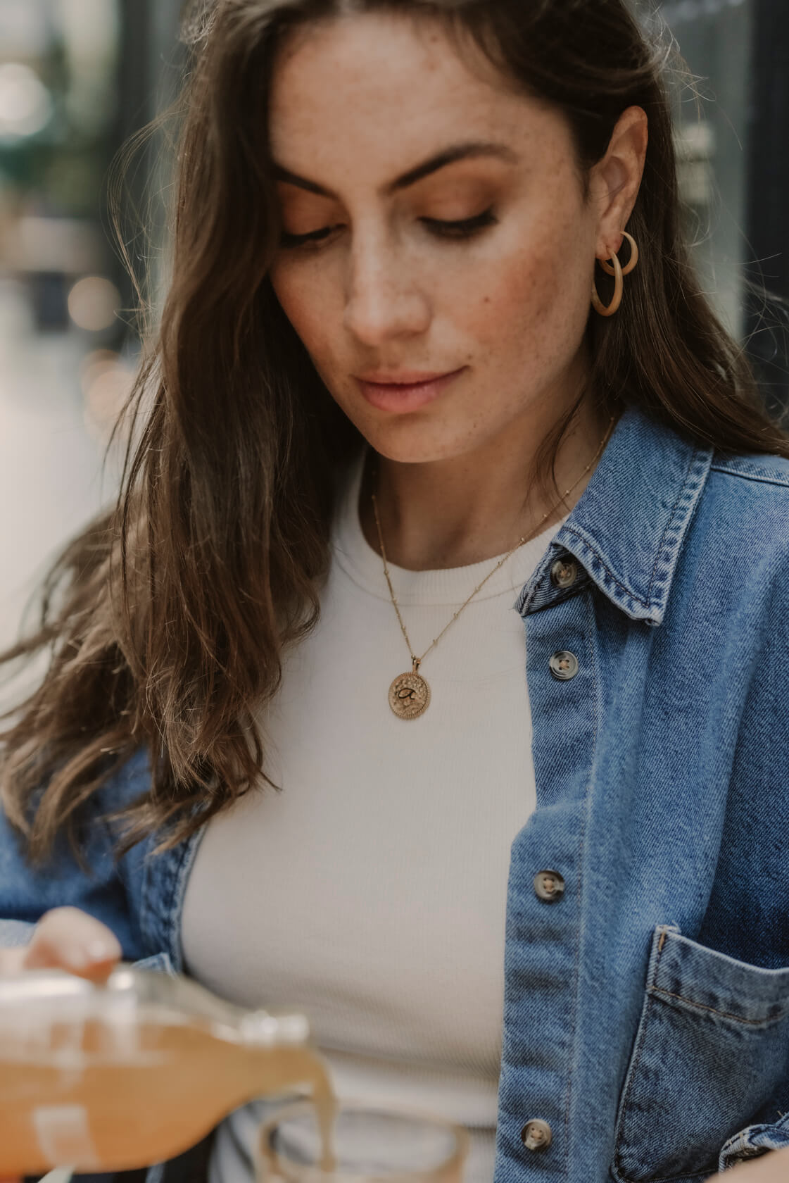 Model is drinking at a cafe wearing large tapered gold hoop earrings and Fuck gold shorthand necklace
