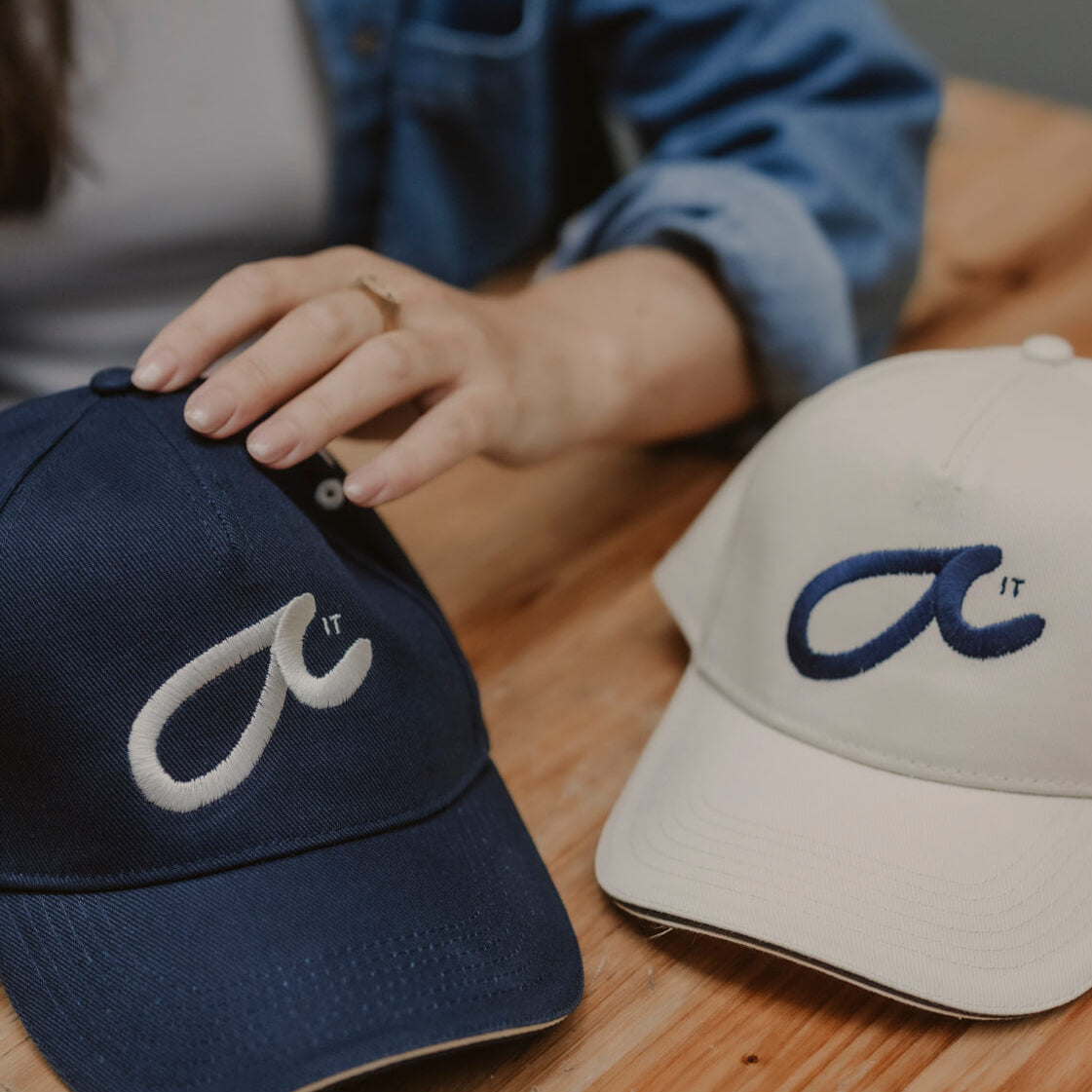 Two baseball caps with logos on a wooden surface, one navy and one putty cream. Logo says Fuck it in Shorthand secret messaging.