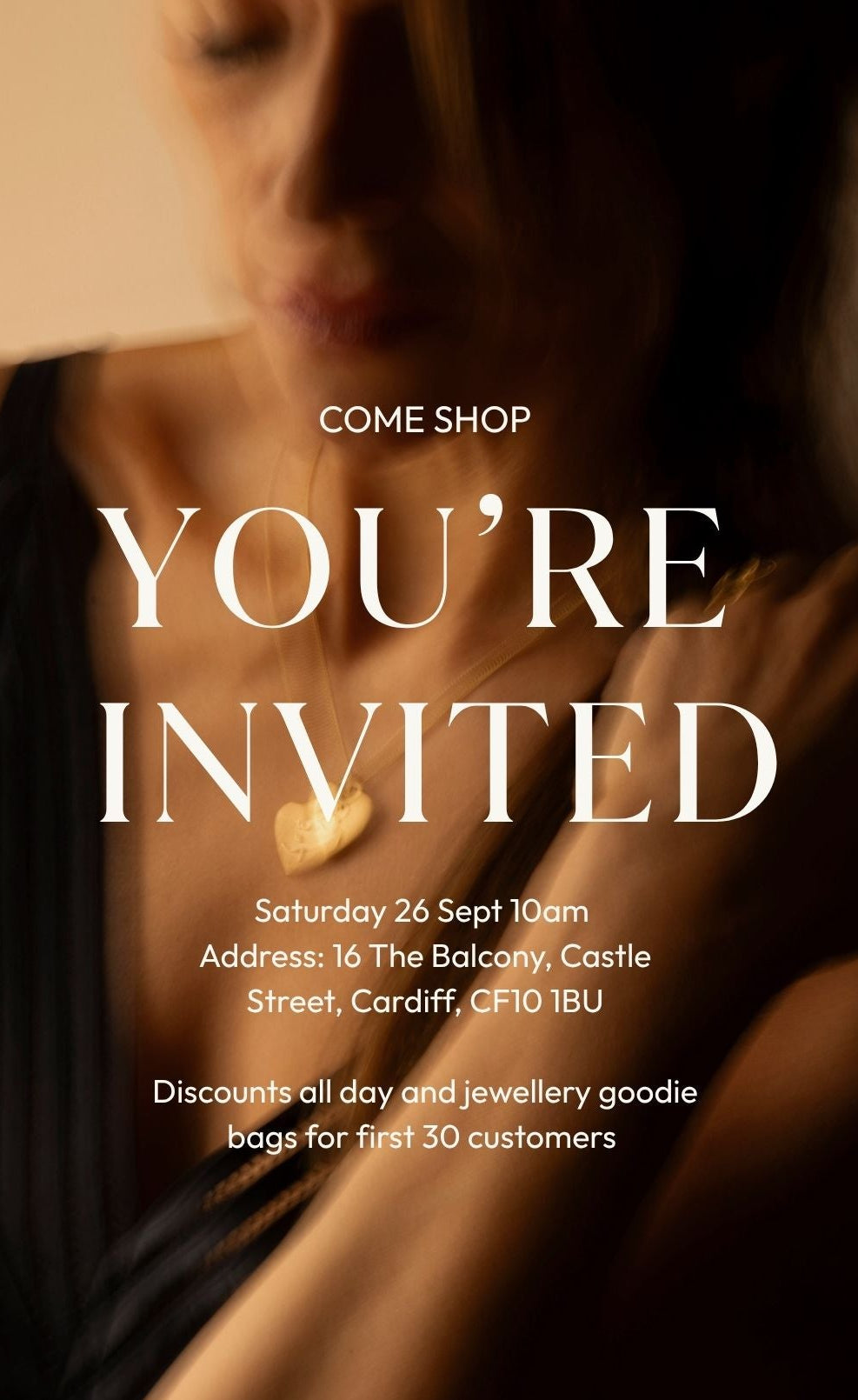Promotional invitation graphic featuring a softly blurred close-up of a woman wearing a black garment and a gold heart-shaped pendant necklace. Large white text reads 'COME SHOP YOU'RE INVITED' with event details below on a dark background.