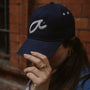 Person wearing a navy blue cap with a white logo which says Fuck It in shorthand against a brick wall.