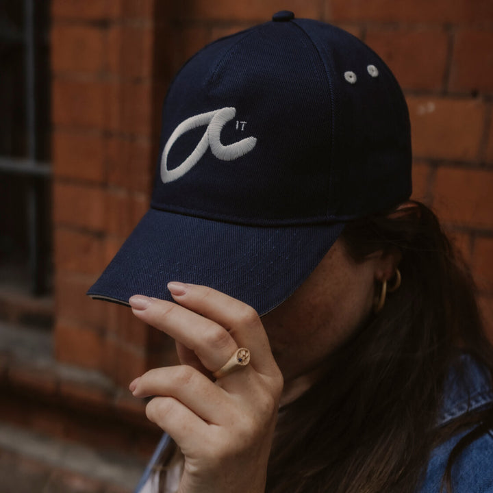 Person wearing a navy blue cap with a white logo which says Fuck It in shorthand against a brick wall.
