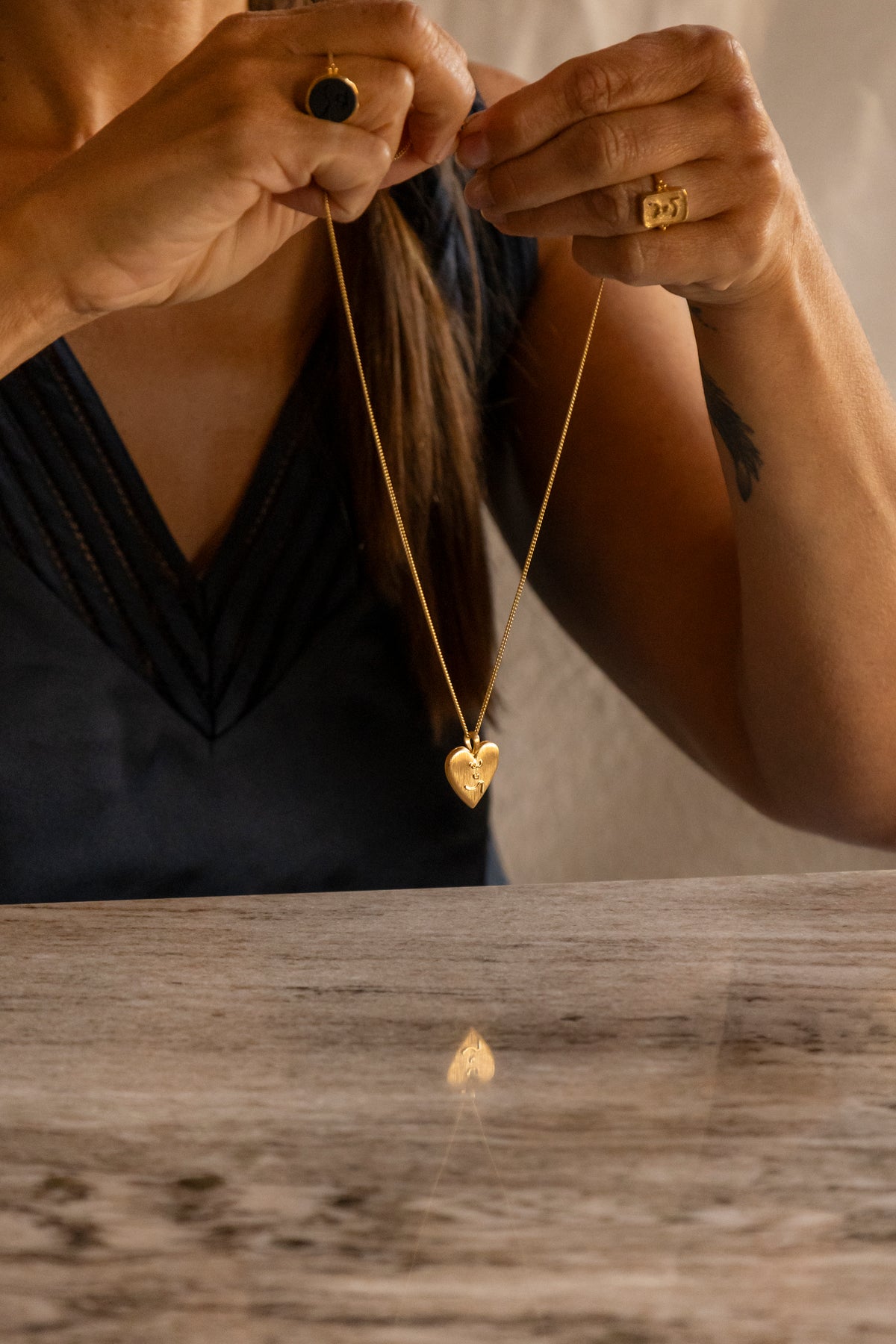 A person holding a gold heart locket pendant with Know Your Worth inscribed in Teeline Shorthand necklace over a wooden table, wearing gold rings on both hands.