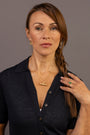 A woman with brown hair in a side braid wears a gold horizontal T bar pendant necklace and a gold ring with a green stone, paired with a dark navy short-sleeve button-up shirt against a solid gray studio background.