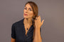 A woman with brown hair in a braid wearing a dark navy short-sleeve polo shirt, a black onyx pendant necklace and matching ring and gold coin huggie earrings all with Difficult Woman in Teeline Shorthand engraved on them
