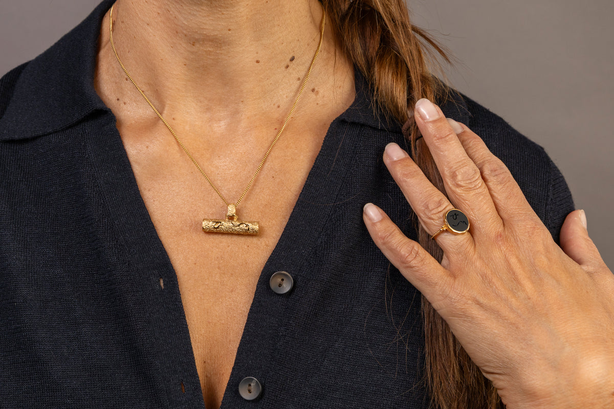A close-up lifestyle shot of a woman wearing a gold T-bar pendant necklace and a gold signet ring with a dark onyx stone both inscribed with Difficult Woman in Teeline Shorthand wearing a dark navy linen shirt.