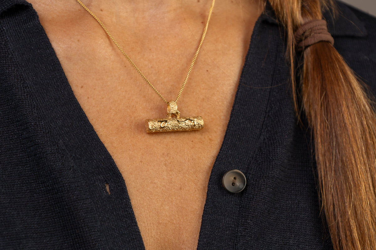 A close-up lifestyle shot of a woman wearing a gold T-bar pendant necklace  inscribed with Difficult Woman in Teeline Shorthand wearing a dark navy linen shirt.