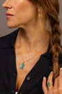 Close-up of a woman wearing a gold chain necklace with a rectangular chalcedony gemstone pendant, along with a matching green ring on her finger and a small gold earring. She is wearing a dark navy button-up shirt.