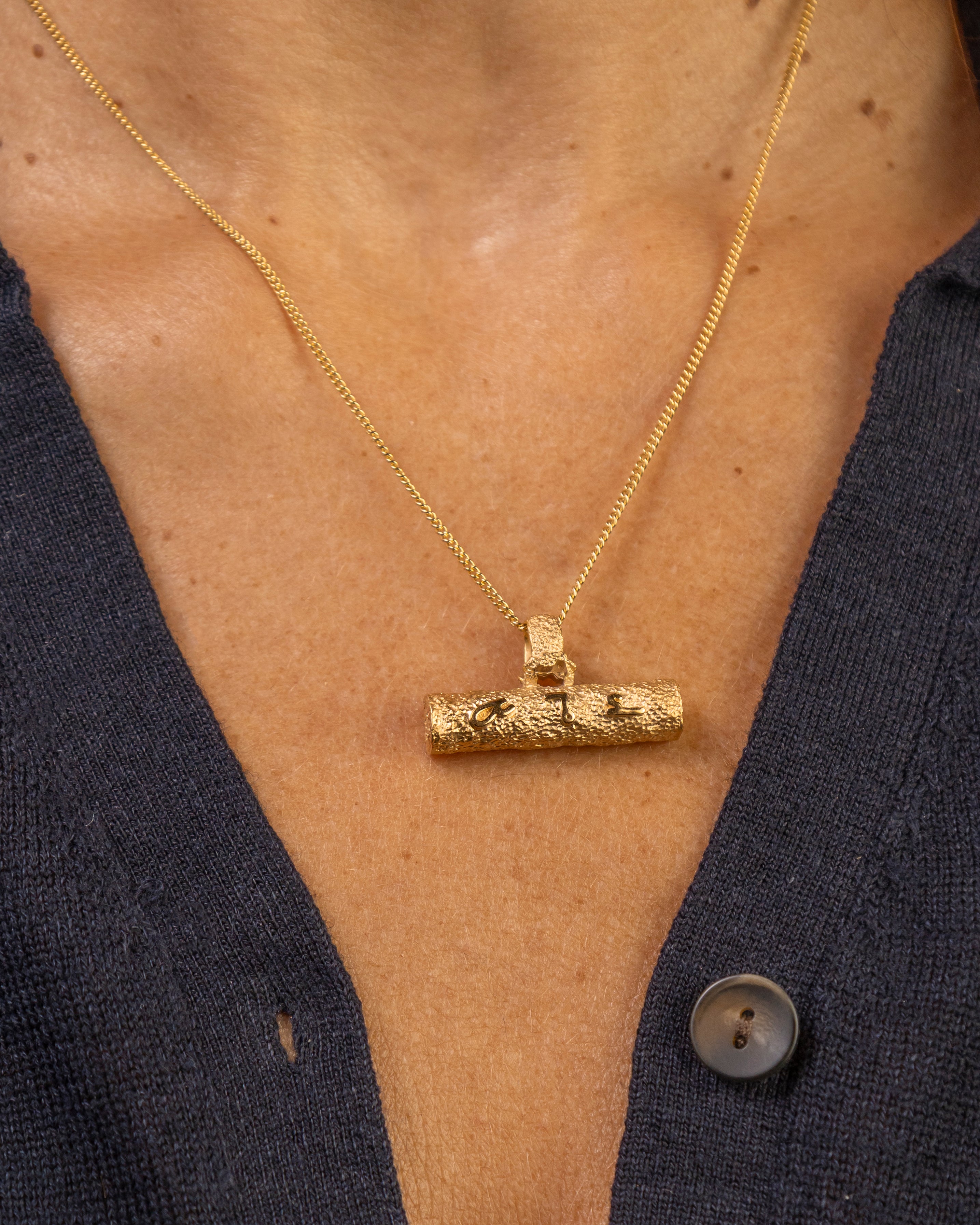 A close-up of a person wearing a textured gold T bar pendant necklace with a F*** this S*** inscribed in Teeline Shorthand, paired with a dark navy button-up shirt.