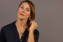 A woman with brown hair in a braid wearing a dark navy short-sleeve polo shirt, a black onyx pendant necklace and matching ring and gold coin huggie earrings all with Difficult Woman in Teeline Shorthand engraved on them