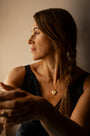 A woman in a black sleeveless top wearing a delicate gold heart pendant locket necklace with Know Your Worth iin Teeline Shorthand, shown in profile against a warm beige background.