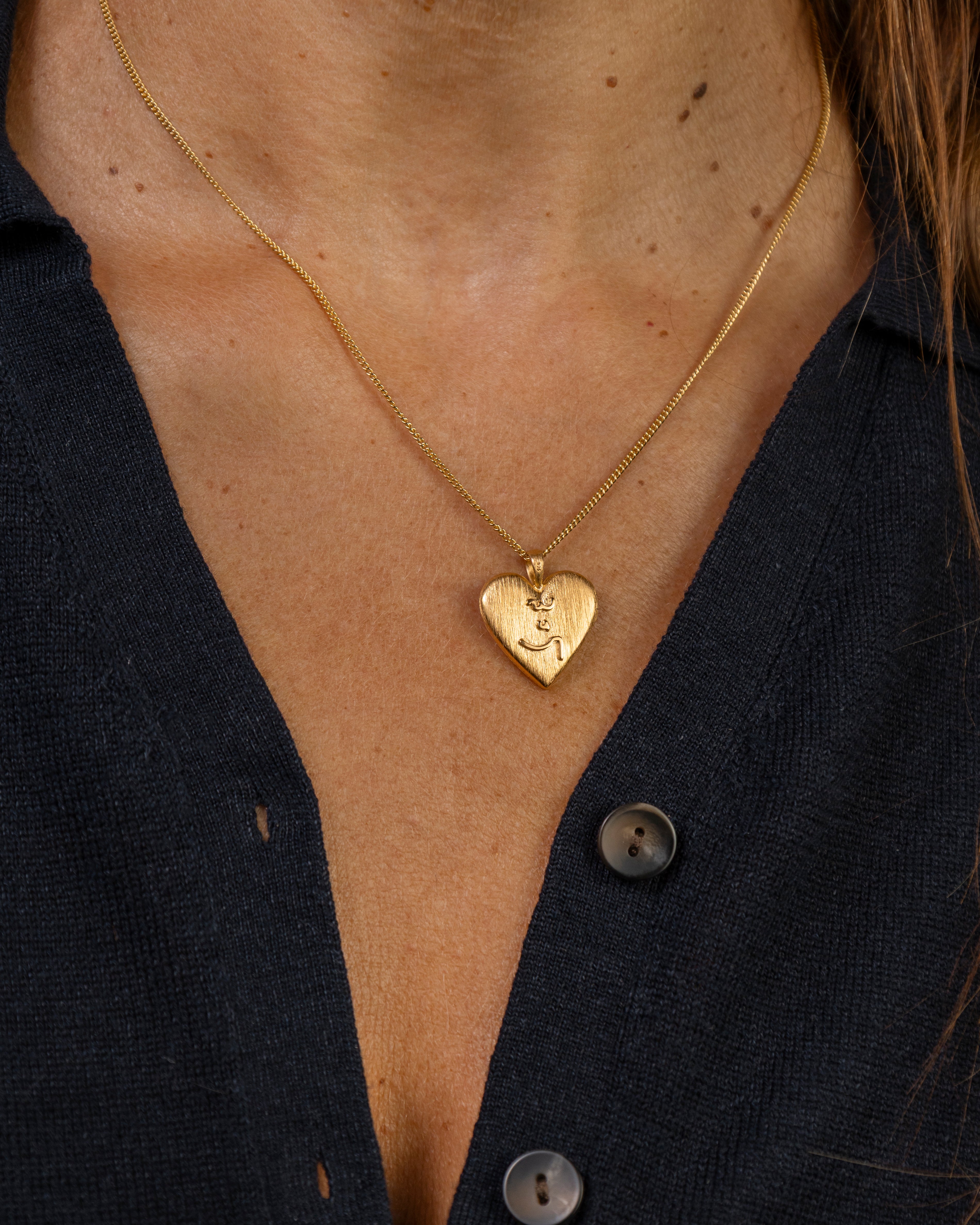A close-up of a person wearing a delicate gold chain necklace with a small heart-shaped pendant engraved with an Know Your Work in Teeline Shorthand symbols, styled with a dark navy button-down shirt.
