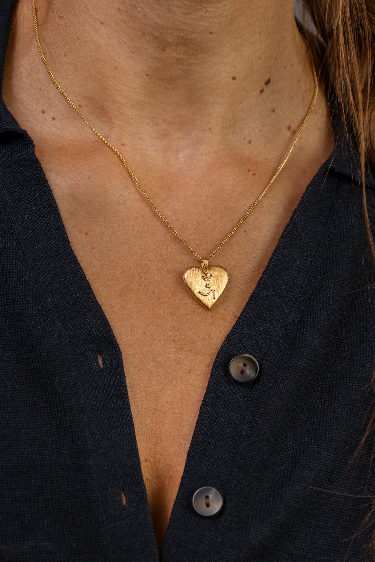 A close-up of a person wearing a delicate gold chain necklace with a small heart-shaped pendant engraved with an Know Your Work in Teeline Shorthand symbols, styled with a dark navy button-down shirt.