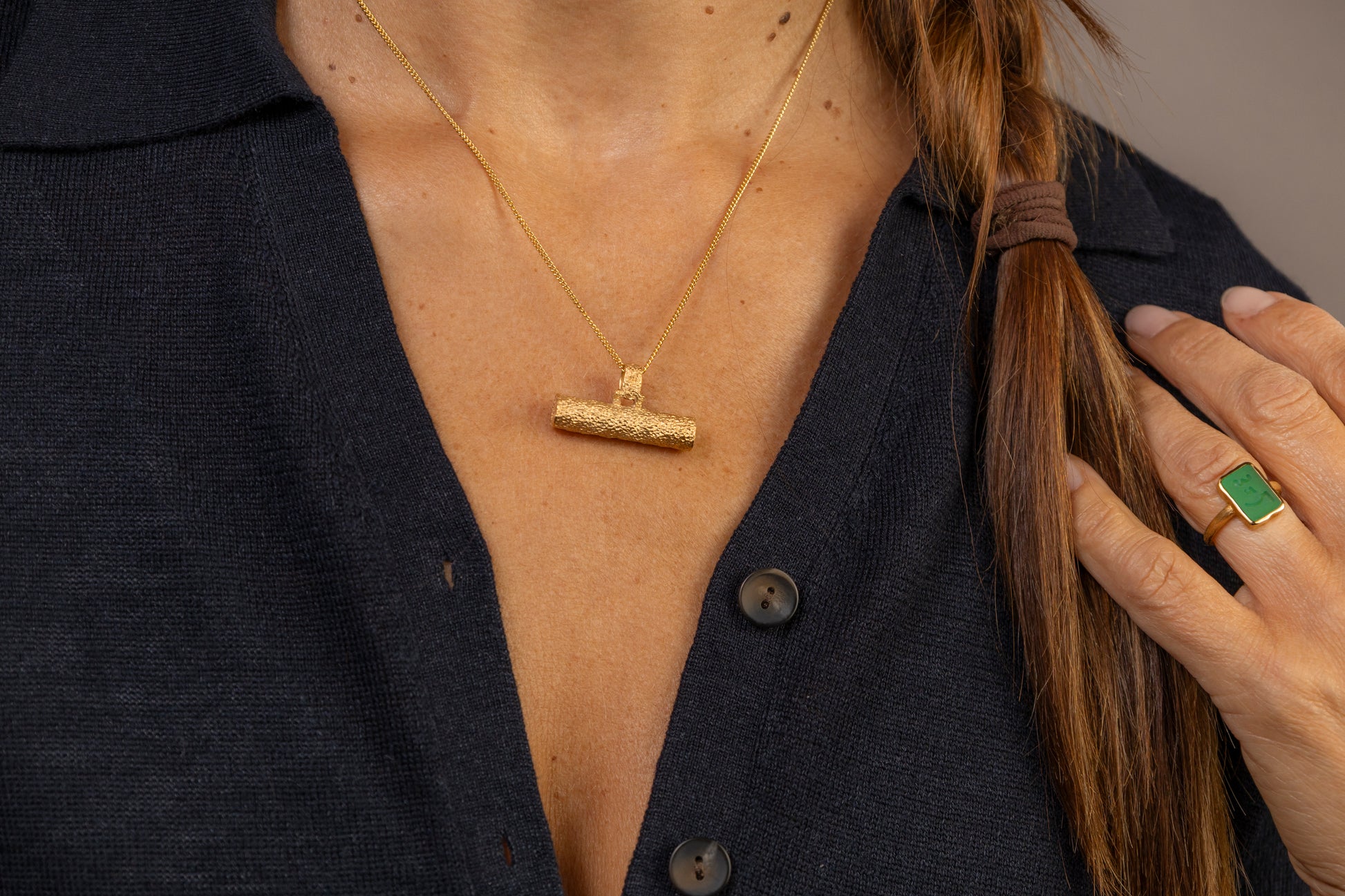 A close-up lifestyle shot of a woman wearing a textured gold T Bar pendant necklace and a gold ring with a green stone, styled with a dark navy knit top.
