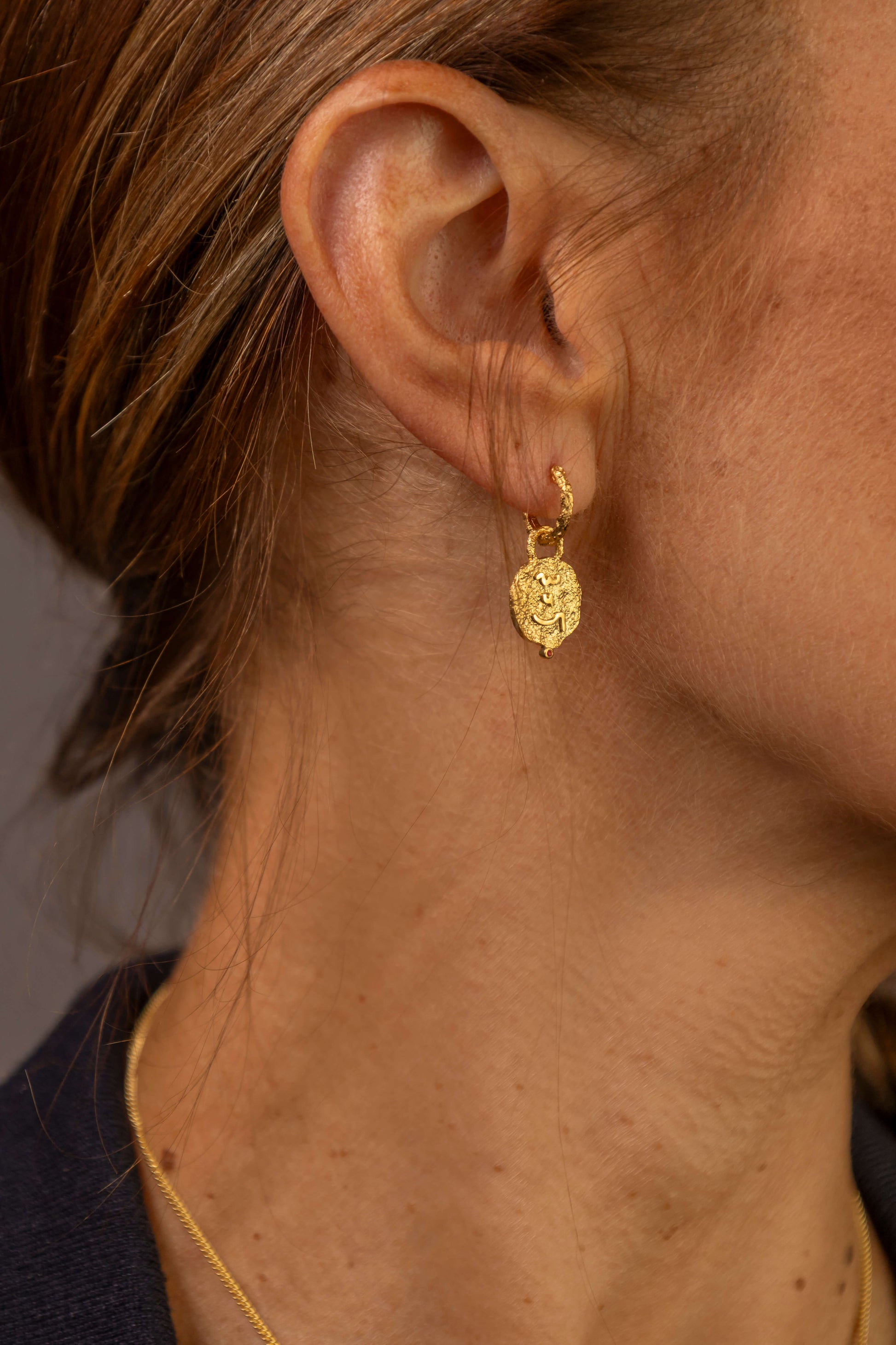 Close up of woman's ear wearing gold textured hoop earrings with coin pendants which say Know Your Worth in Shorthand 