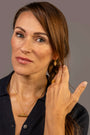 A woman with brown hair in a braid wearing gold jewelry, including a gold necklace, ring, and earring, against a solid gray background. The T Bar nekclace, hoop earrings and ring all say Know Your Worth in Teeline Shorthand symbols