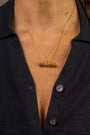 A close-up of a person wearing a gold necklace with a horizontal T bar pendant gold necklace with Know Your Worth in Teeline Shorthand, paired with a dark navy button-up shirt.