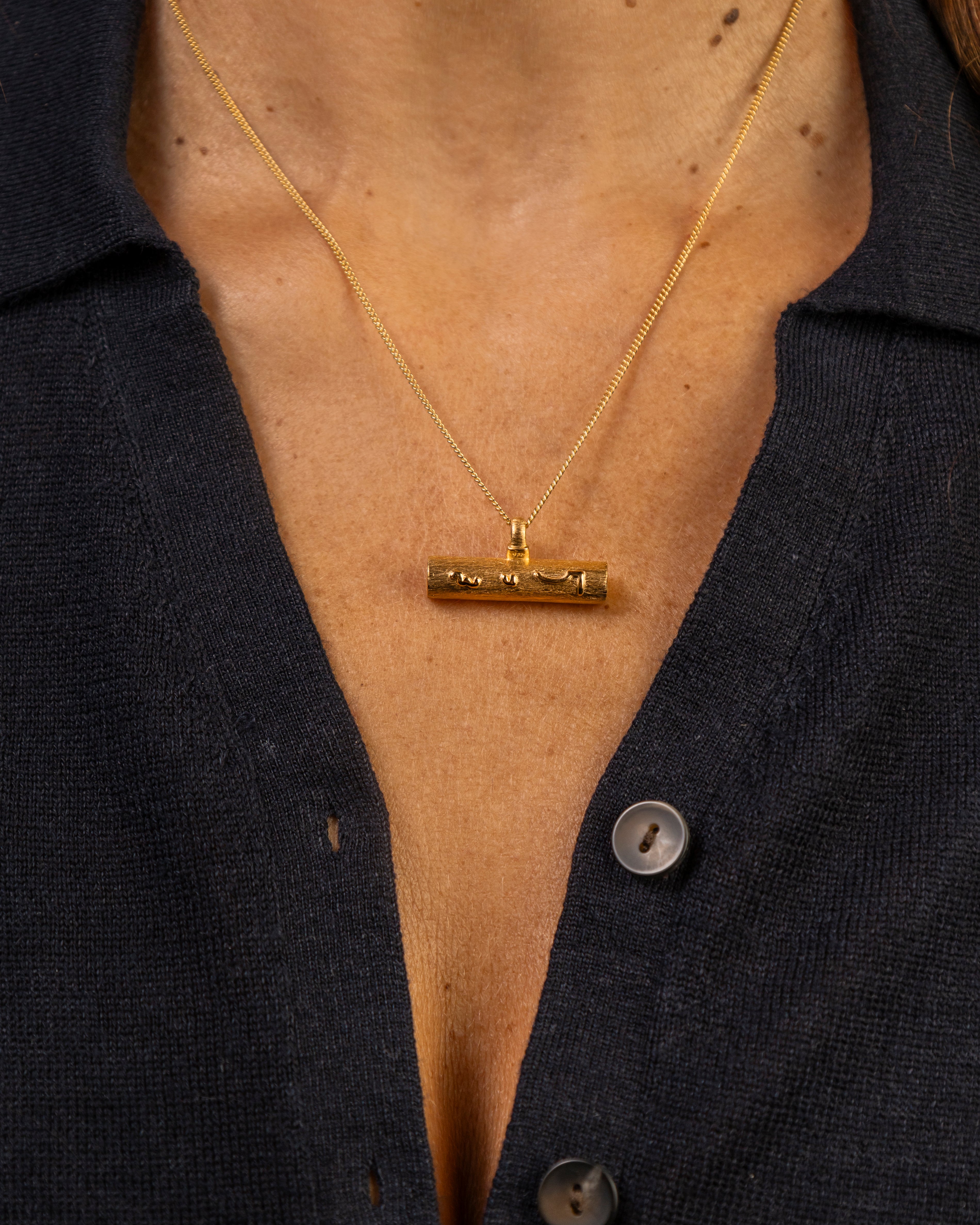 A close-up of a person wearing a gold necklace with a horizontal T bar pendant gold necklace with Know Your Worth in Teeline Shorthand, paired with a dark navy button-up shirt.
