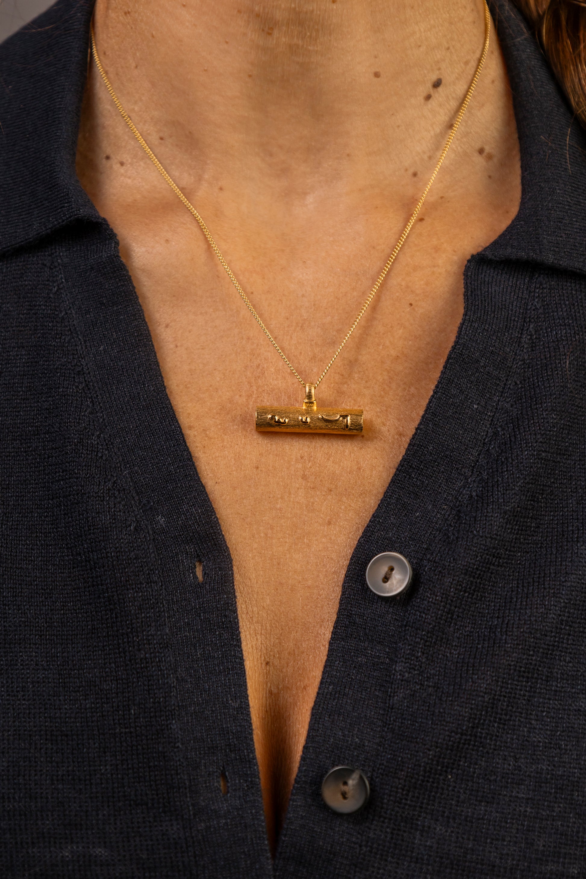 A close-up of a person wearing a gold necklace with a horizontal T bar pendant gold necklace with Know Your Worth in Teeline Shorthand, paired with a dark navy button-up shirt.