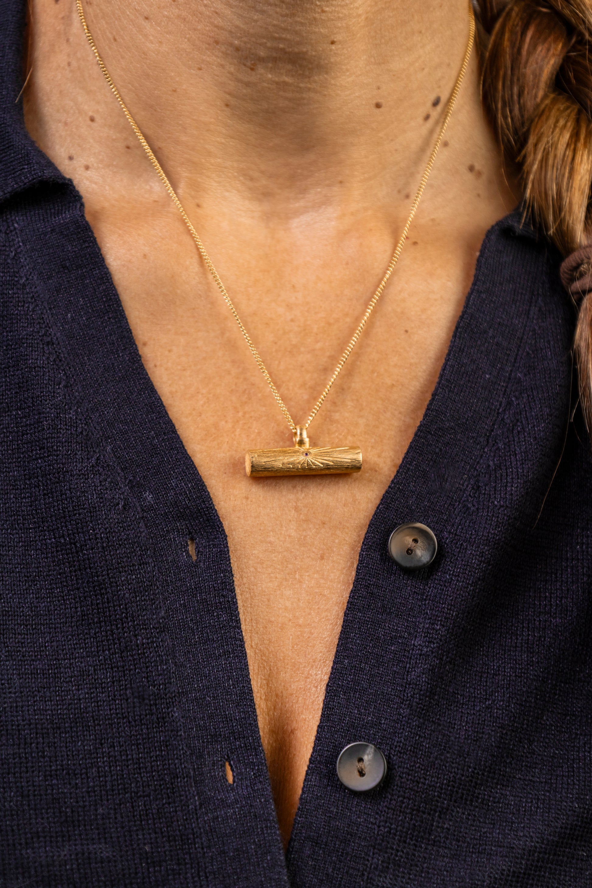 A close-up of a person wearing a gold necklace with a horizontal T bar pendant gold necklace with ruby gemstone in Teeline Shorthand, paired with a dark navy button-up shirt.