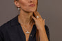 A close-up of a woman wearing a silver necklace with a silver T-bar pendant inscribed with F*** this S*** in Teeline Shorthand and matching silver hoop earrings, shown against a solid gray background.