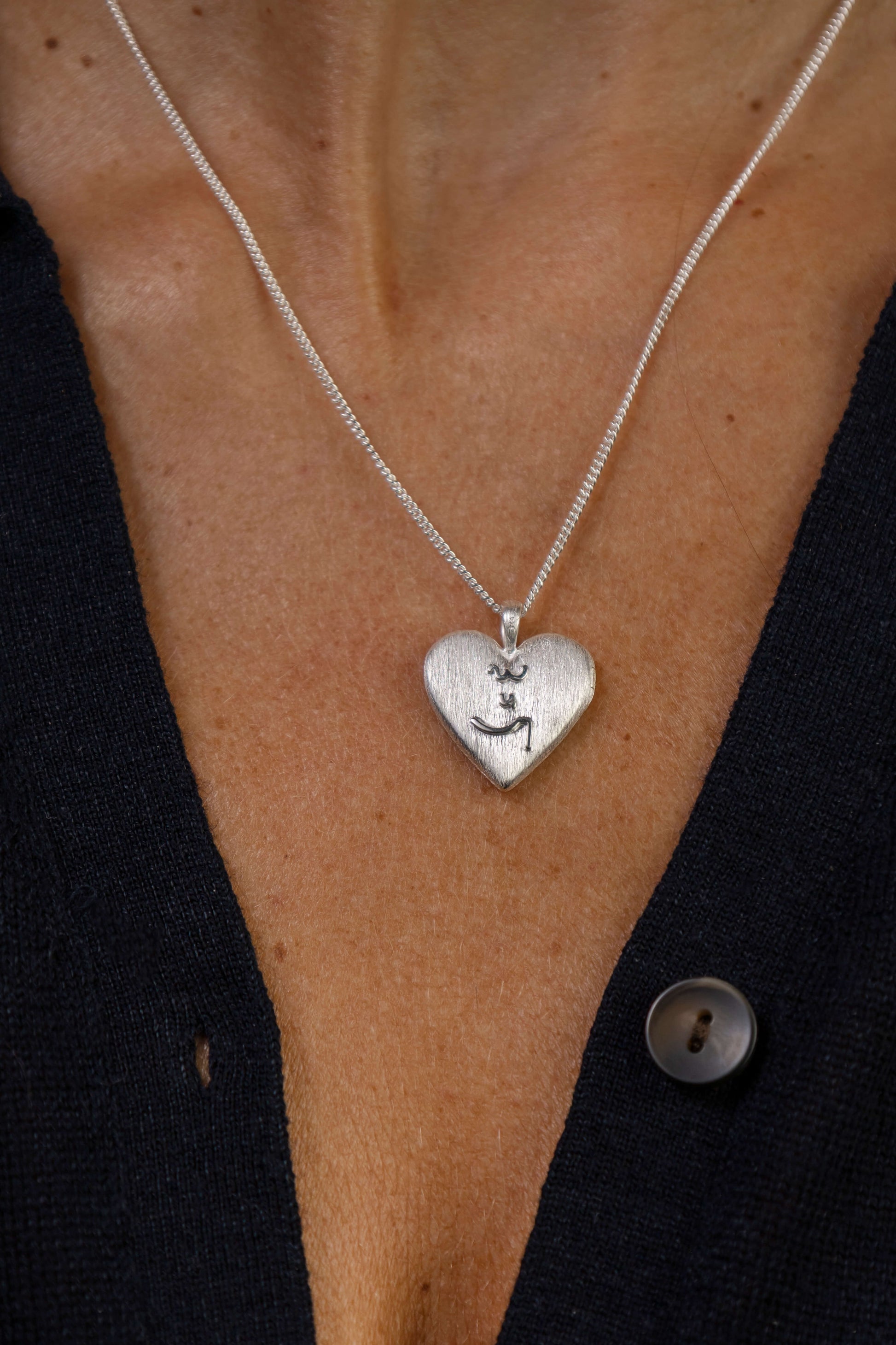A close-up of a person wearing a delicate silver chain necklace with a textured heart-shaped silver locket with Know Your Worth in Teeline Shorthand in the middle. 