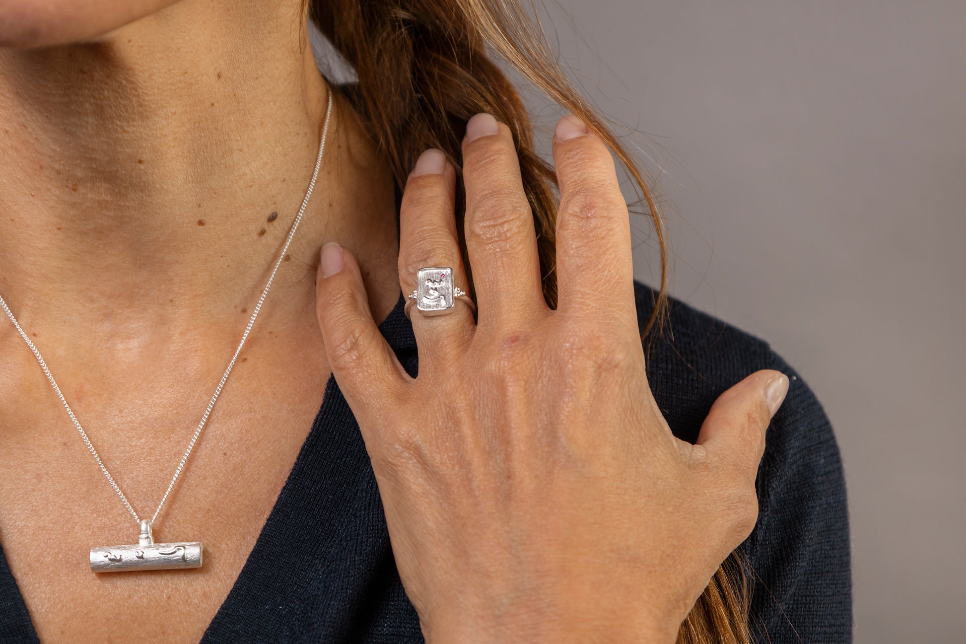 A woman with brown hair is shown from the chest up against a solid gray studio background, wearing a silver T  bar pendant necklace and a silver ring with Know Your Worth inscribed on both  in Teeline Shorthand 
