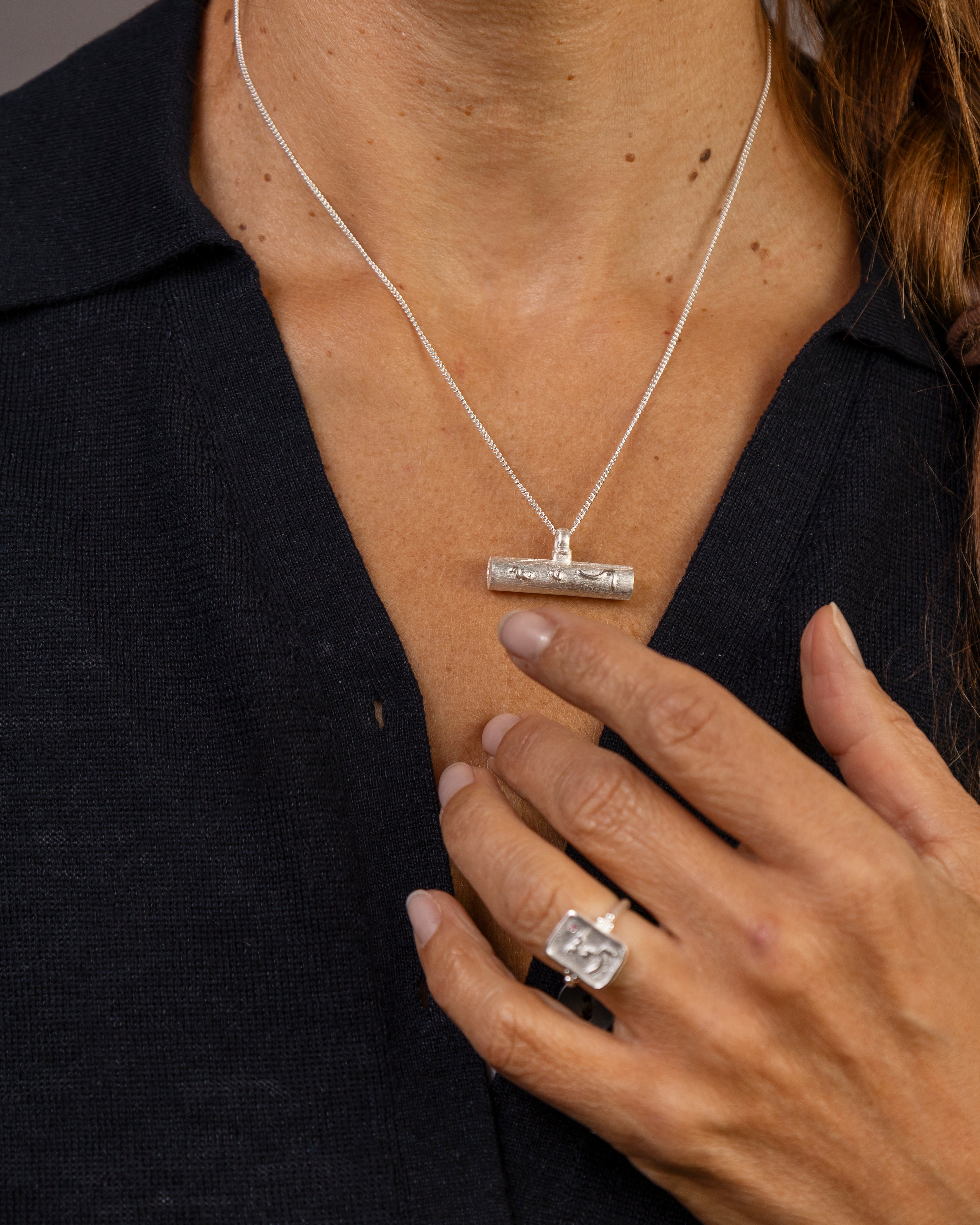 A woman with brown hair is shown from the chest up against a solid gray studio background, wearing a silver T  bar pendant necklace and a silver ring with Know Your Worth inscribed on both  in Teeline Shorthand 
