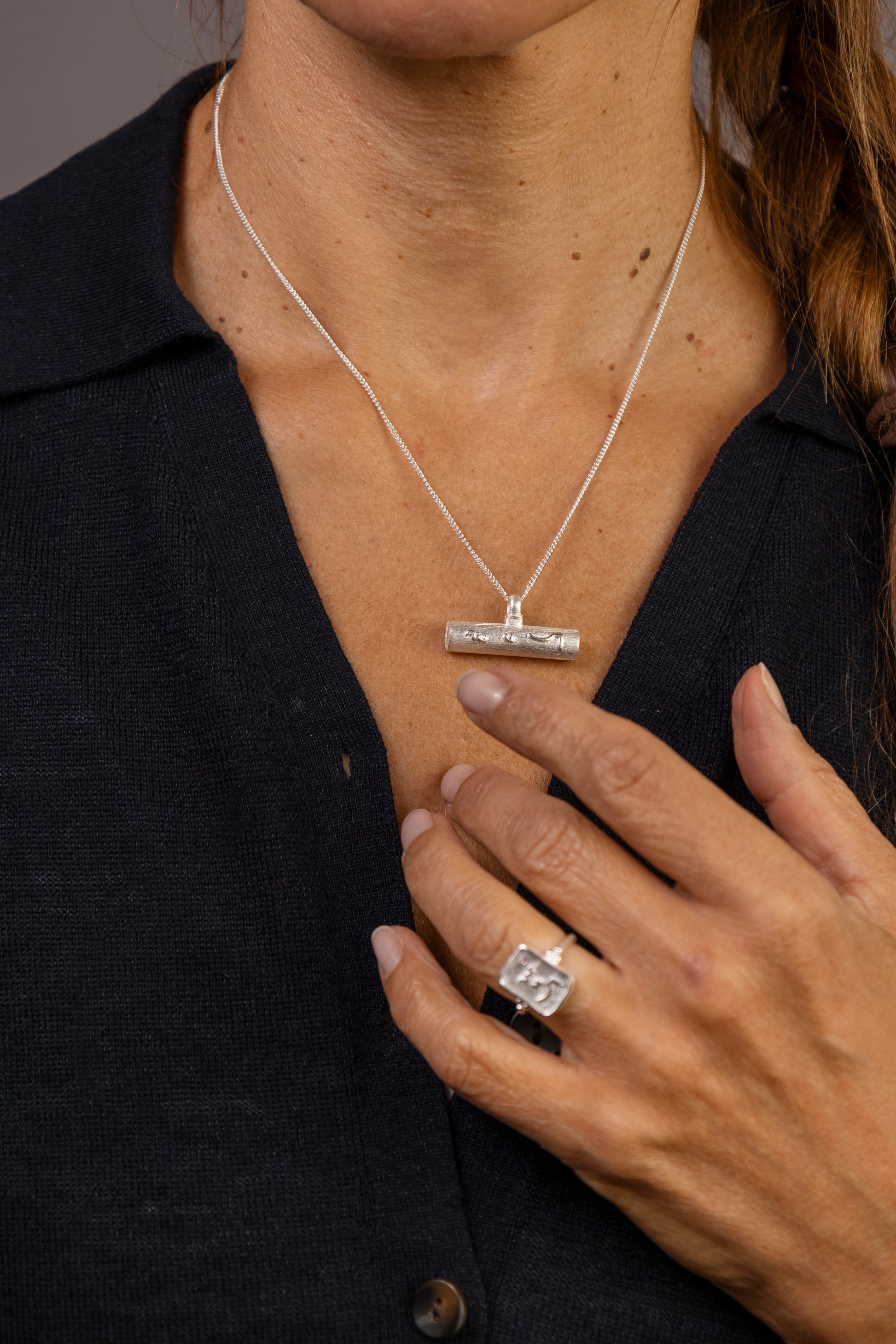 A woman with brown hair is shown from the chest up against a solid gray studio background, wearing a silver T  bar pendant necklace and a silver ring with Know Your Worth inscribed on both  in Teeline Shorthand 
