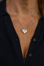 A close-up of a person wearing a delicate silver chain necklace with a textured heart-shaped silver locket with a sacred heart and ruby gemstone in the middle. 
