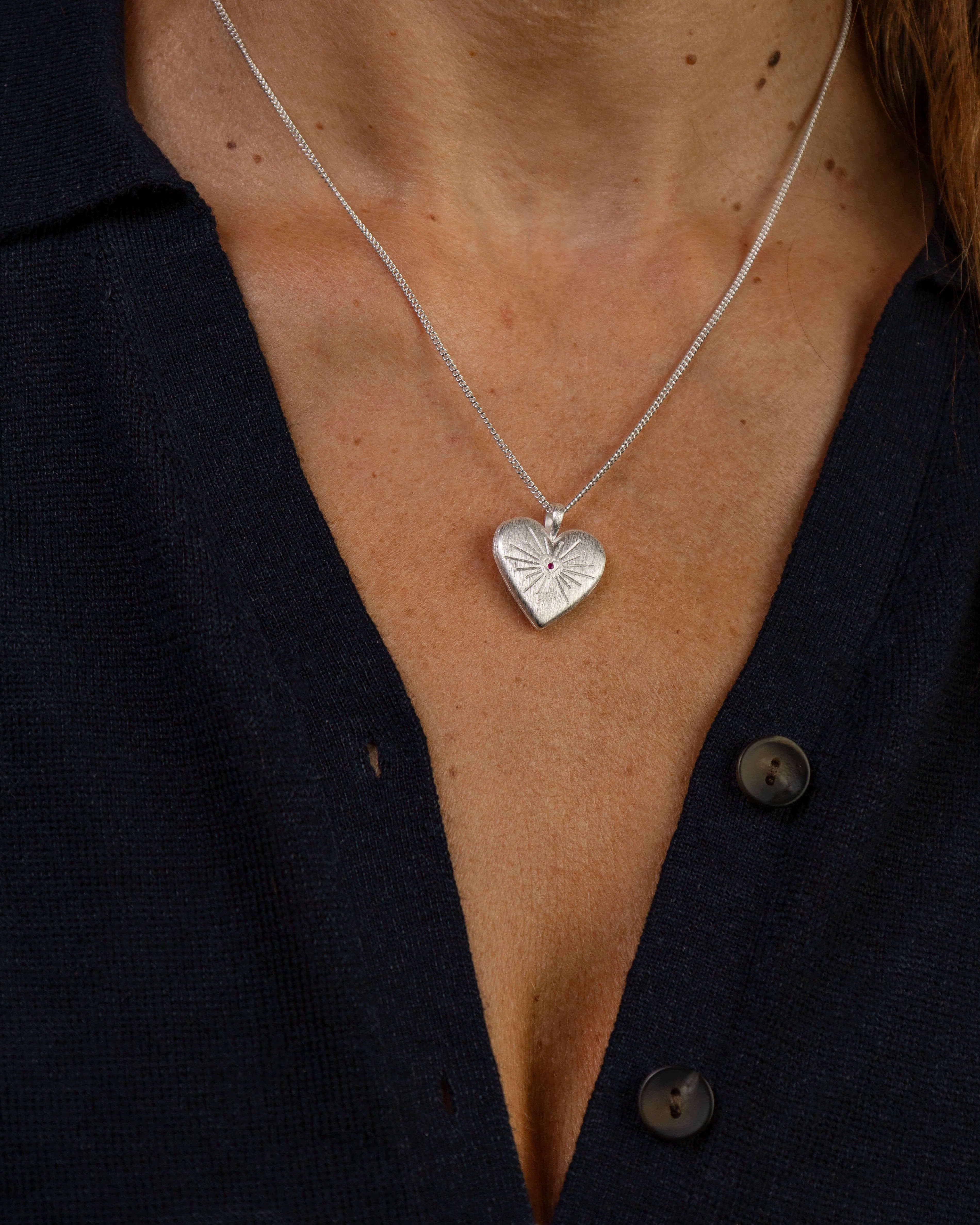 A close-up of a person wearing a delicate silver chain necklace with a textured heart-shaped silver locket with a sacred heart and ruby gemstone in the middle. 
