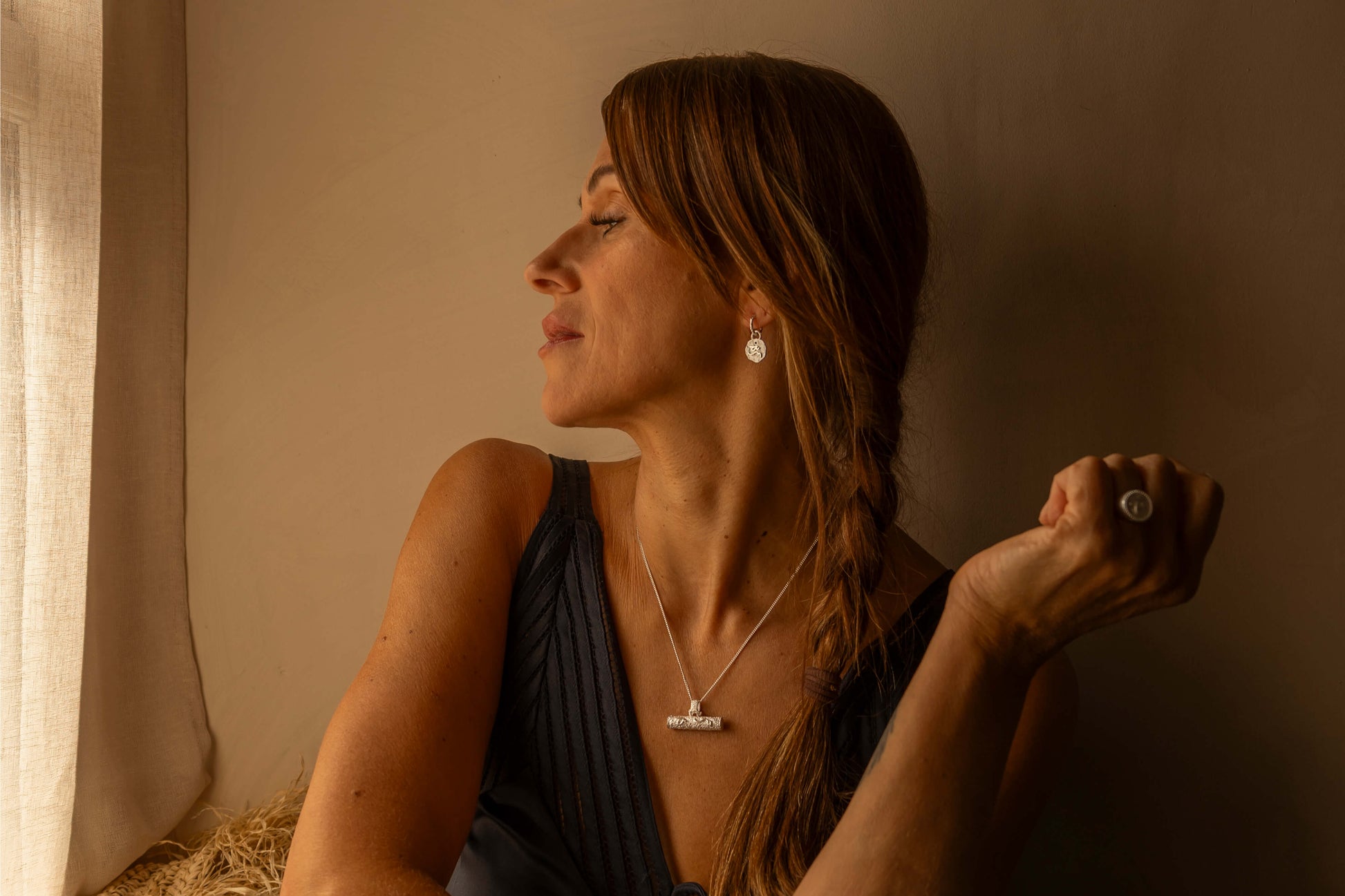 A woman with long brown hair in a side braid is shown in profile, wearing a silver T bar pendant necklace and a silver ring inscribed with Difficult Woman in Teeline Shorthand, set against a warm, dimly lit beige wall.