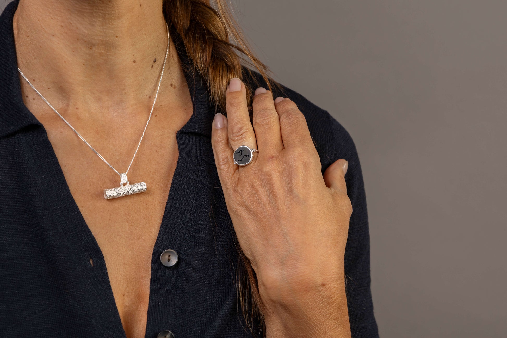 A close-up of a person wearing a textured silver reversible T Bar Necklace necklace and a silver signet ring with a black Onyx gemstone with Difficult Woman in Teeline Shorthand symbols inscribed into the stone, paired with a dark navy button-up shirt against a solid gray background.